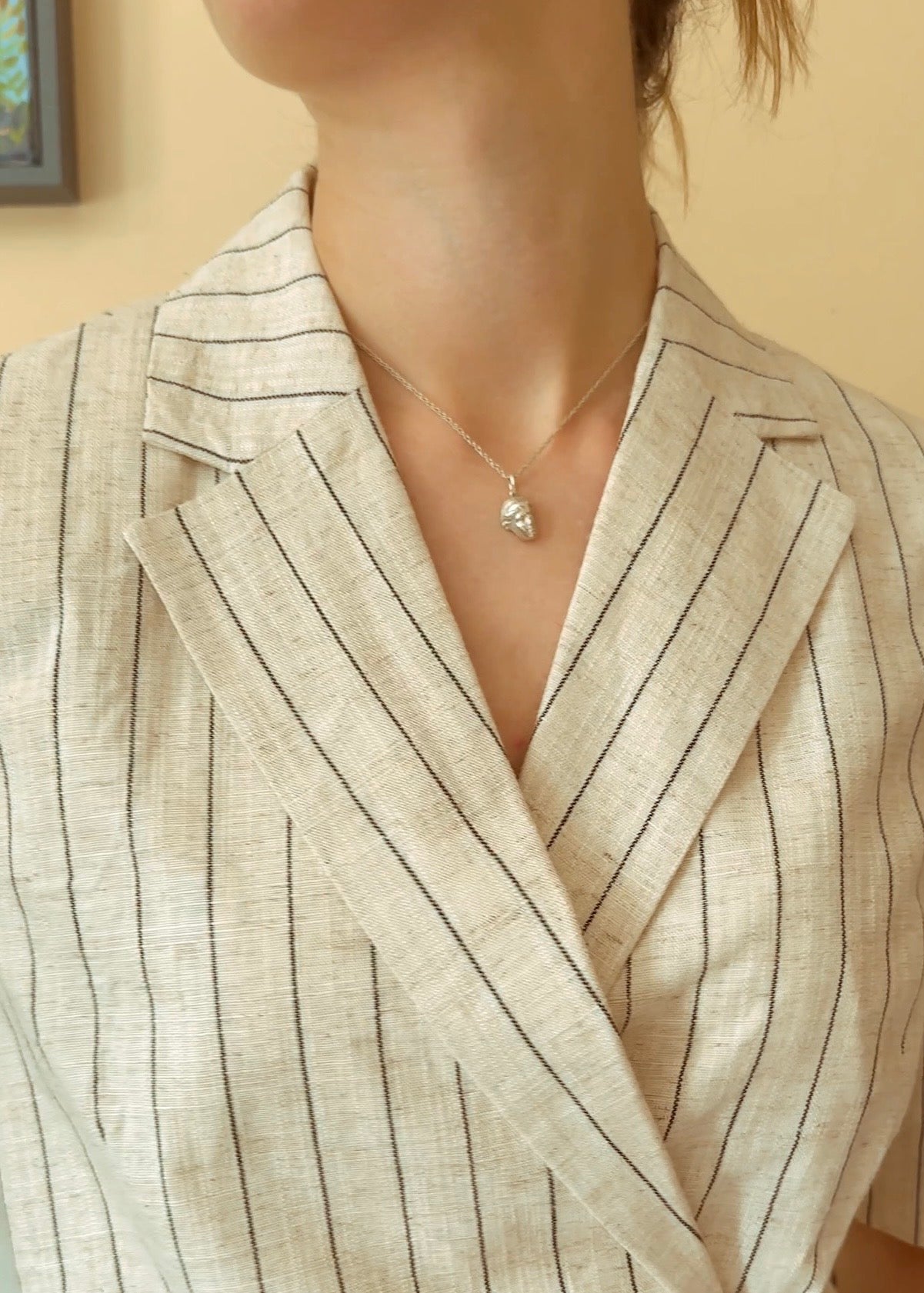 A close-up of a person wearing a delicate silver pendant necklace of Venus de Milo's face, styled with a beige pinstriped linen blazer.